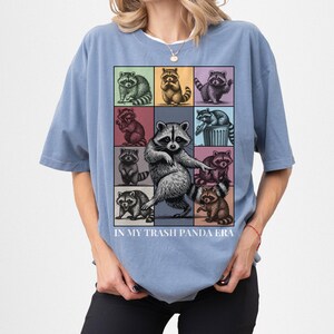 May include: Blue t-shirt with a grid of raccoon illustrations in various poses and colors. The text "IN MY TRASH PANDA ERA" is printed below the grid. The shirt is a solid blue color.