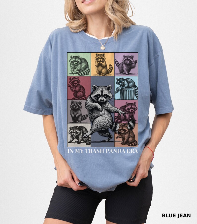 May include: Blue t-shirt with a grid of raccoon illustrations in various poses and colors. The text "IN MY TRASH PANDA ERA" is printed below the grid. The shirt is a solid blue color.