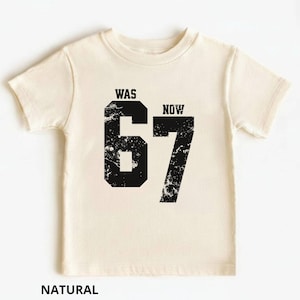 Peut inclure: Un t-shirt &agrave; manches courtes de couleur naturelle avec un grand graphique noir repr&eacute;sentant le nombre 67 dans une texture vieillie. Au-dessus du chiffre 6, le mot WAS est imprim&eacute;, et au-dessus du chiffre 7, le mot NOW est imprim&eacute; en noir.