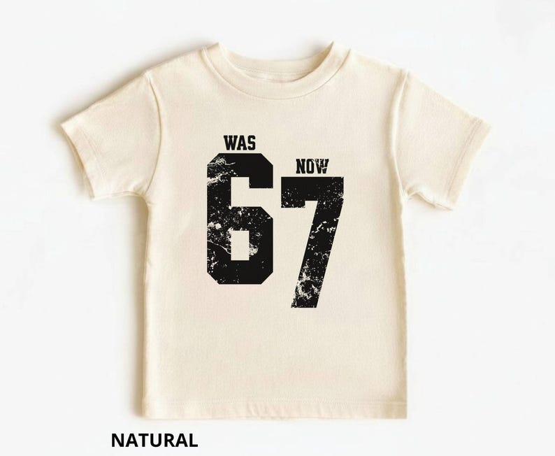 Peut inclure: Un t-shirt &agrave; manches courtes de couleur naturelle avec un grand graphique noir repr&eacute;sentant le nombre 67 dans une texture vieillie. Au-dessus du chiffre 6, le mot WAS est imprim&eacute;, et au-dessus du chiffre 7, le mot NOW est imprim&eacute; en noir.