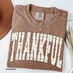 May include: Espresso-colored t-shirt with the word "THANKFUL" in large, white, block letters filled with a leaf pattern. A silver necklace with a pendant and a brown hat are also visible. The shirt is labeled "COMFORT COLORS".