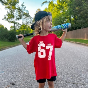 Peut inclure: Un t-shirt rouge &agrave; manches courtes avec le texte blanc vieilli WAS 67 NOW. Le v&ecirc;tement est port&eacute; avec une casquette de baseball noire et un short noir, tandis qu'une batte de baseball &agrave; motifs bleus et blancs est tenue sur les &eacute;paules.