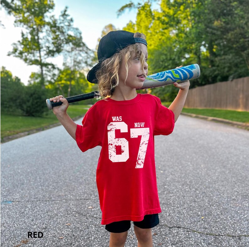 Peut inclure: Un t-shirt rouge &agrave; manches courtes avec le texte blanc vieilli WAS 67 NOW. Le v&ecirc;tement est port&eacute; avec une casquette de baseball noire et un short noir, tandis qu'une batte de baseball &agrave; motifs bleus et blancs est tenue sur les &eacute;paules.