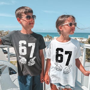 Puede incluir: Dos niños con camisetas con el número "67" en blanco y negro desgastado. Las camisetas también presentan un gráfico de dos manos señalando. Una camiseta es gris oscuro, la otra es blanca. Ambos niños llevan gafas de sol.