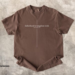 May include: Espresso-colored t-shirt with the text "fatherhood is kingdom work" and "PROVERBS 22:6" in white. A vertical white line is centered below the text. The shirt is made of a soft material.