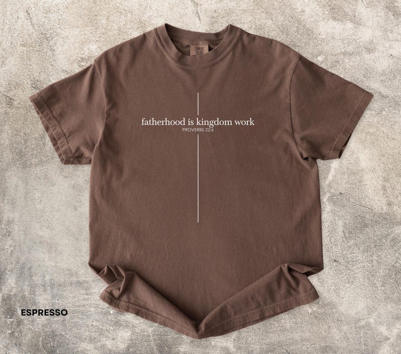 May include: Espresso-colored t-shirt with the text "fatherhood is kingdom work" and "PROVERBS 22:6" in white. A vertical white line is centered below the text. The shirt is made of a soft material.