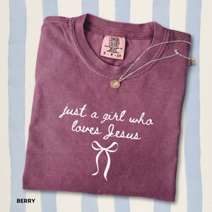 May include: A berry-colored short-sleeve t-shirt with a crew neckline and a Comfort Colors brand label. The front features white text that reads "just a girl who loves Jesus" above a white graphic of a bow. A thin silver-tone necklace rests on top.