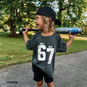 Peut inclure: Un t-shirt gris anthracite &agrave; manches courtes avec le texte blanc vieilli WAS 67 NOW 67, associ&eacute; &agrave; une casquette de baseball noire et un short noir. Le t-shirt est port&eacute; en tenant une batte de baseball argent&eacute;e et bleue sur les &eacute;paules.