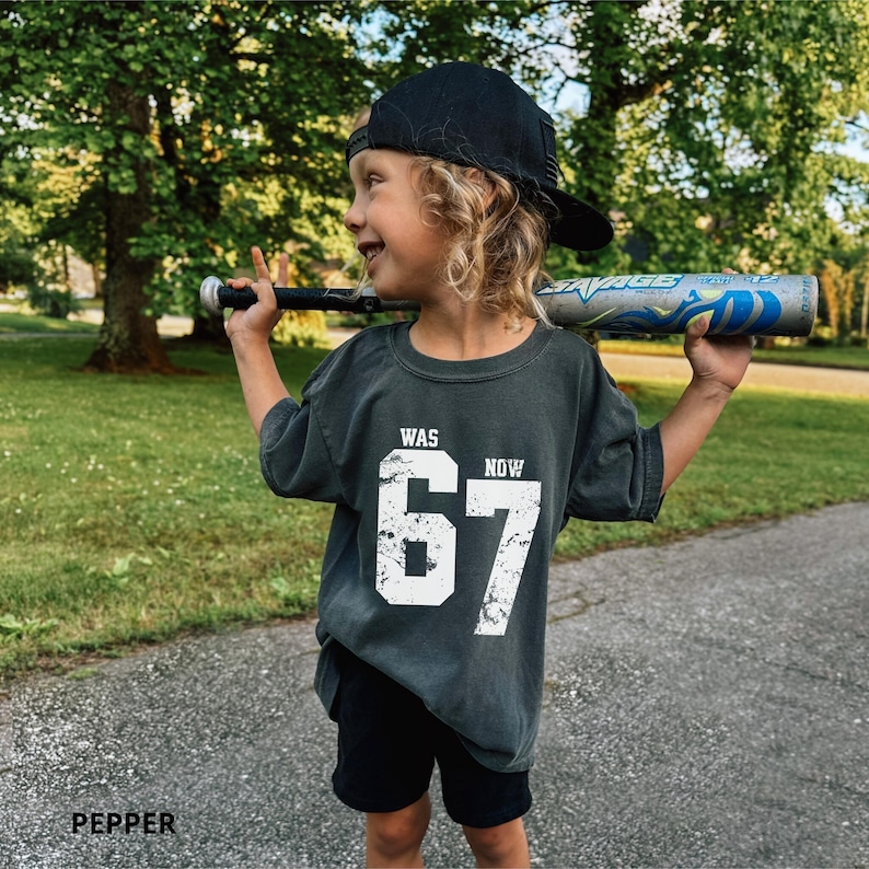 Peut inclure: Un t-shirt gris anthracite &agrave; manches courtes avec le texte blanc vieilli WAS 67 NOW 67, associ&eacute; &agrave; une casquette de baseball noire et un short noir. Le t-shirt est port&eacute; en tenant une batte de baseball argent&eacute;e et bleue sur les &eacute;paules.