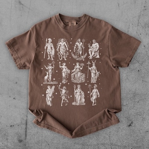 May include: Brown t-shirt featuring a white illustration of various classical figures. The figures are arranged in rows and columns, with stars scattered around them. The shirt has a crew neck and short sleeves.