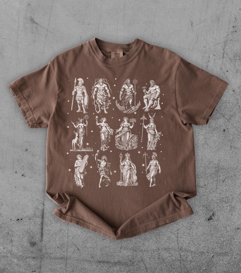 May include: Brown t-shirt featuring a white illustration of various classical figures. The figures are arranged in rows and columns, with stars scattered around them. The shirt has a crew neck and short sleeves.