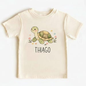 Puede incluir: Camiseta color crema con una tortuga de dibujos animados con caparazón verde y marrón, rodeada de pequeñas flores. El nombre "THIAGO" está impreso en negro debajo. La camiseta es de un material suave.
