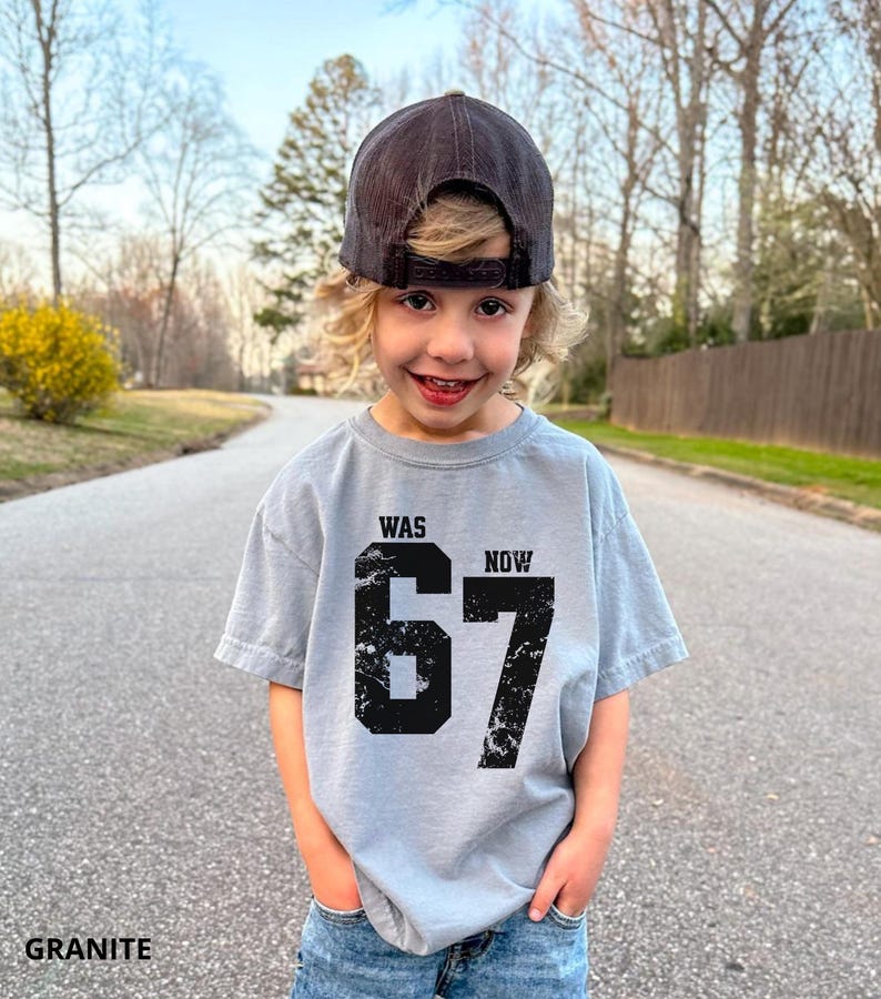 Peut inclure: Un t-shirt gris clair &agrave; manches courtes avec une impression graphique noire vieillie. Le motif comprend le mot WAS au-dessus du nombre 67 et le mot NOW &agrave; droite du nombre. Le t-shirt est port&eacute; avec une casquette de baseball gris fonc&eacute;.