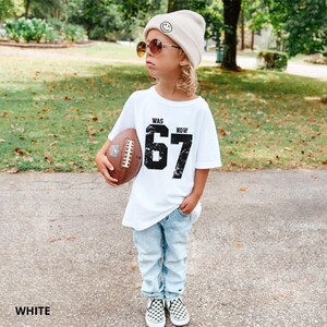 Peut inclure: Un t-shirt blanc avec le texte noir WAS 67 NOW pr&eacute;sentant un effet graphique vieilli. Le haut est associ&eacute; &agrave; un jean en denim bleu clair, un bonnet beige avec une ic&ocirc;ne de visage souriant, des lunettes teint&eacute;es marron et un ballon de football.