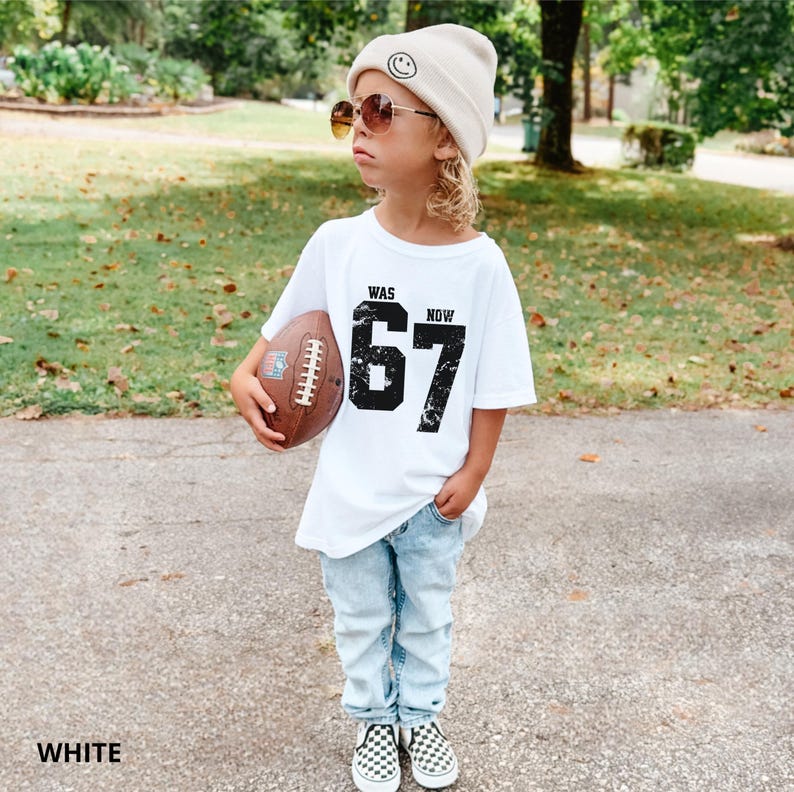 Peut inclure: Un t-shirt blanc avec le texte noir WAS 67 NOW pr&eacute;sentant un effet graphique vieilli. Le haut est associ&eacute; &agrave; un jean en denim bleu clair, un bonnet beige avec une ic&ocirc;ne de visage souriant, des lunettes teint&eacute;es marron et un ballon de football.