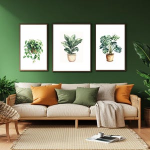 Peut inclure: Trois impressions d'art mural encadrées représentant des illustrations à l'aquarelle de plantes en pot. L'impression de gauche montre une plante grimpante, celle du centre une plante à larges feuilles en pot beige, et celle de droite un monstera.