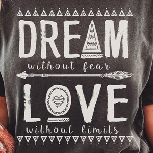 May include: Dark gray t-shirt with the words "DREAM without fear LOVE without limits" in white. The design includes arrow and geometric shapes. The shirt is worn with denim shorts and a patterned blanket.