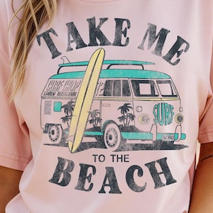 May include: A light pink t-shirt with a graphic of a vintage van carrying a surfboard, palm trees, and the words "TAKE ME TO THE BEACH" in black. The shirt is a relaxed fit.