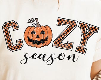 May include: Cream-colored t-shirt with the words "COZY season" in black script and orange block letters. The letter "C" and "Z" have a black and orange checkered pattern. A smiling jack-o'-lantern is in the center.