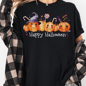 May include: Black t-shirt with a Halloween-themed graphic. The design features smiling orange pumpkins wearing a witch hat and holding candy canes, with the words "Happy Halloween" in white.