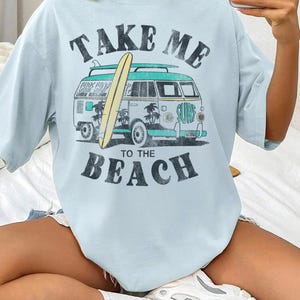 May include: Light blue t-shirt with a vintage-style graphic. The design features a teal and white van with a surfboard, palm trees, and the words "TAKE ME TO THE BEACH" in a distressed font. The shirt has short sleeves.