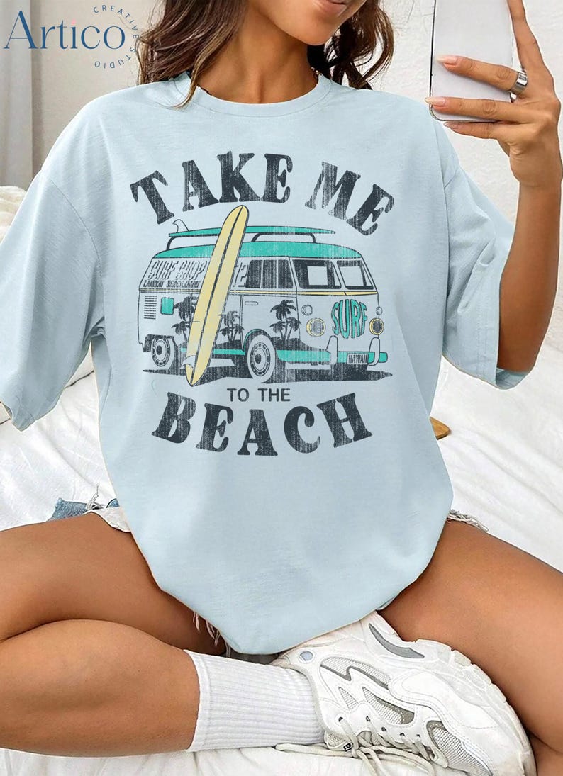 May include: Light blue t-shirt with a vintage-style graphic. The design features a teal and white van with a surfboard, palm trees, and the words "TAKE ME TO THE BEACH" in a distressed font. The shirt has short sleeves.