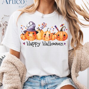 May include: White t-shirt with a colorful Halloween design. The graphic features smiling orange pumpkins wearing a witch hat and holding candy. The text "Happy Halloween" is printed below the pumpkins, with small purple hearts.