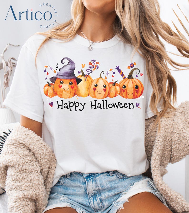 May include: White t-shirt with a colorful Halloween design. The graphic features smiling orange pumpkins wearing a witch hat and holding candy. The text "Happy Halloween" is printed below the pumpkins, with small purple hearts.