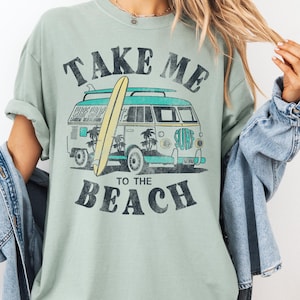 May include: Sage green t-shirt with a vintage graphic of a surf van and surfboard. The text on the shirt reads "TAKE ME TO THE BEACH" in a distressed font. The van is teal and white, with palm trees and the word "SURF" on the side.