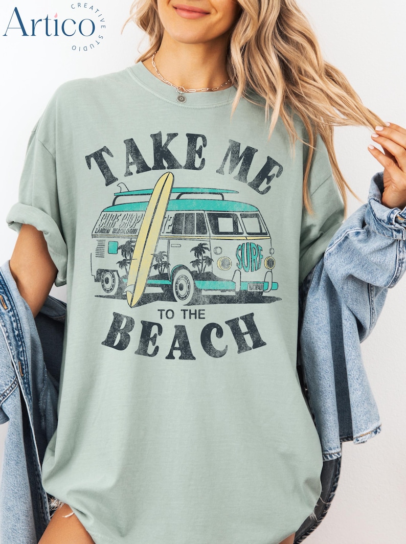 May include: Sage green t-shirt with a vintage graphic of a surf van and surfboard. The text on the shirt reads "TAKE ME TO THE BEACH" in a distressed font. The van is teal and white, with palm trees and the word "SURF" on the side.