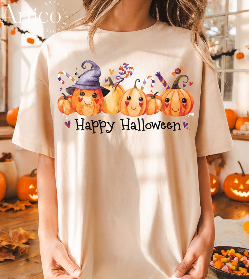 May include: Beige t-shirt featuring a watercolor illustration of smiling pumpkins wearing a witch hat and surrounded by candy. The text "Happy Halloween" is printed below the pumpkins. The shirt is a relaxed fit.