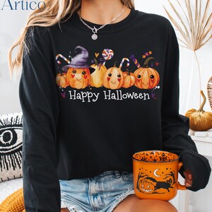 May include: Black long-sleeve shirt with a "Happy Halloween" graphic. The graphic features smiling pumpkins, candy, and a witch's hat. An orange Halloween-themed mug is held in the hand.