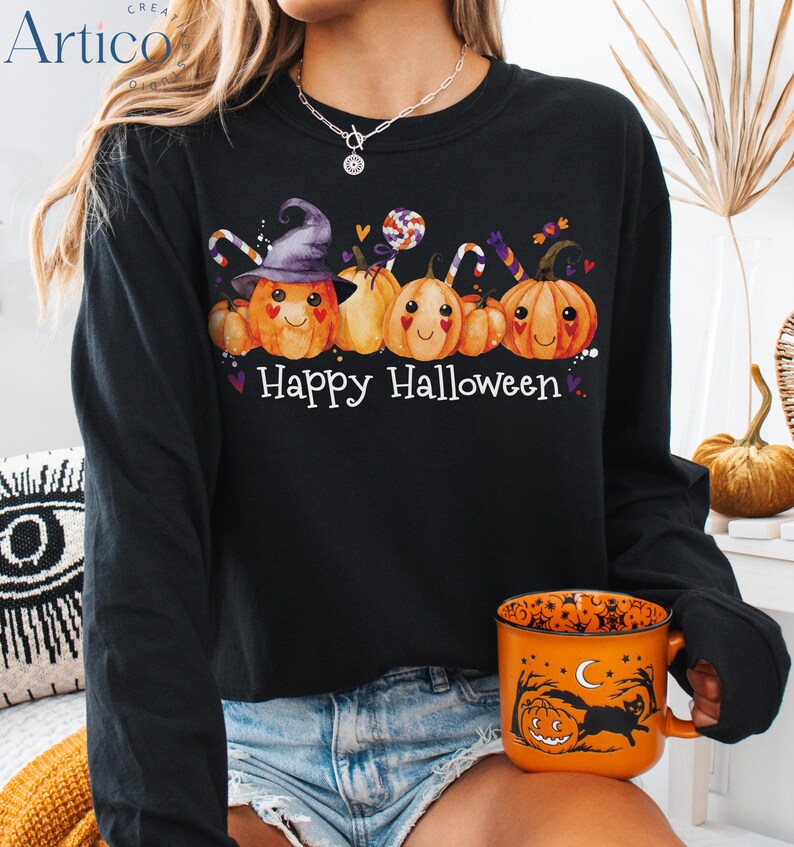May include: Black long-sleeve shirt with a "Happy Halloween" graphic. The graphic features smiling pumpkins, candy, and a witch's hat. An orange Halloween-themed mug is held in the hand.