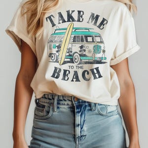 May include: Cream-colored t-shirt with a retro surf design. The graphic features a vintage van with a surfboard, palm trees, and the words "TAKE ME TO THE BEACH" in black. The shirt has rolled-up sleeves.