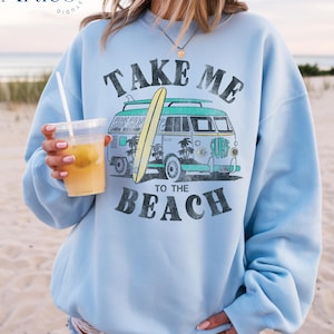 May include: Light blue sweatshirt with a graphic of a vintage van carrying a surfboard, palm trees, and the text "TAKE ME TO THE BEACH". The sweatshirt is worn on a beach.