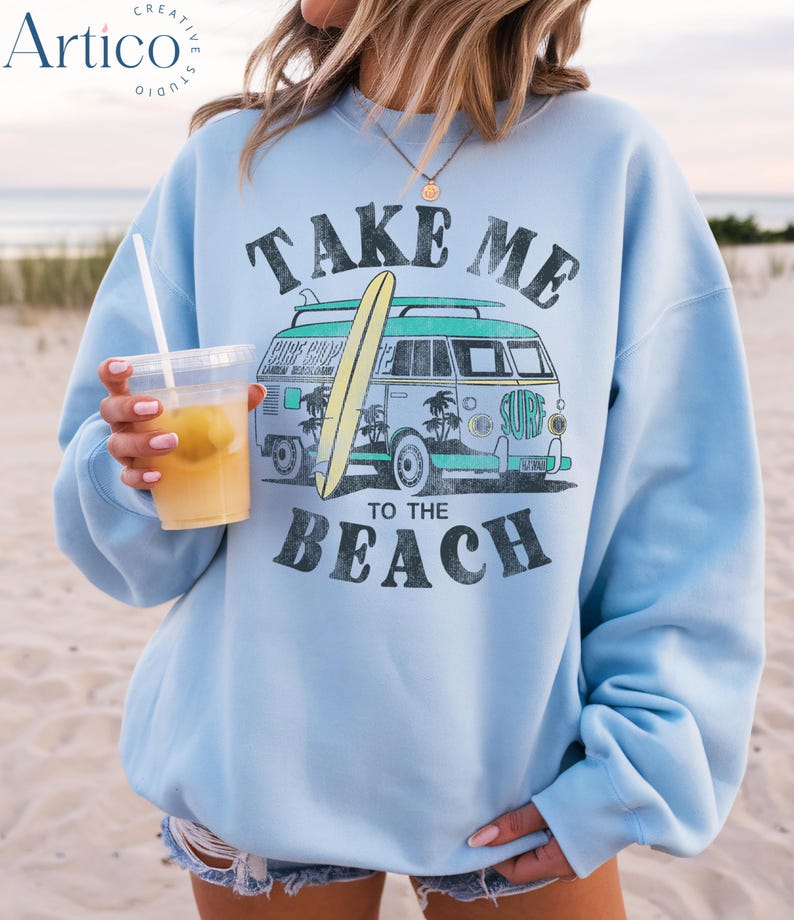 May include: Light blue sweatshirt with a graphic of a vintage van carrying a surfboard, palm trees, and the text "TAKE ME TO THE BEACH". The sweatshirt is worn on a beach.