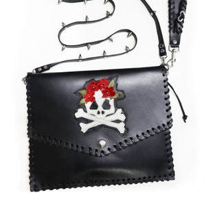 May include: Black leather shoulder bag with a skull and crossbones design and red flowers. The bag has a studded strap and a zipper closure.