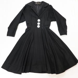 May include: A solid black long-sleeved dress featuring a wide collar, a fitted bodice, and a flared skirt. The front of the garment is secured with three large, circular white buttons positioned vertically along the center of the waistline.