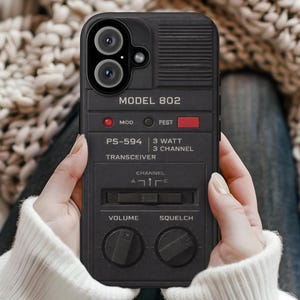 May include: Black phone case with a retro transceiver design. The case features the text "MODEL 802", "PS-594", and "TRANSCEIVER". It has volume and squelch knobs, and a channel slider. The case is held in front of a cozy, neutral-toned background.