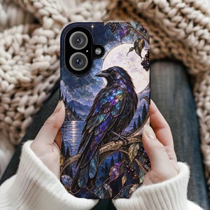 May include: A phone case featuring a detailed illustration of a black raven with iridescent purple and blue feathers perched on a branch. The background depicts a full moon over a dark lake, mountains, and forest, rendered in a stained glass style.