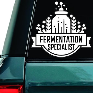 Puede incluir: Calcomanía de vinilo blanco con la frase "Fermentation Specialist" en una pancarta, acompañada de una ilustración estilizada de un recipiente de fermentación y elementos circundantes. La calcomanía está en la ventana trasera de un vehículo verde azulado.