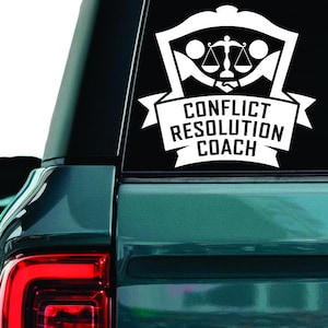 Può includere: Decalcomania bianca sul lunotto posteriore di un veicolo. La decalcomania presenta un disegno a scudo con bilance della giustizia e la scritta "Conflict Resolution Coach" in uno striscione sottostante. Il veicolo è verde acqua.