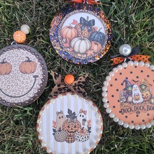 May include: Four circular Halloween ornaments featuring festive designs: one with a leopard print smiley face and pumpkins, one with a pumpkin patch, one with striped ghosts and pumpkins, and one with ducks and ghosts labeled DUCK, DUCK, BOO.