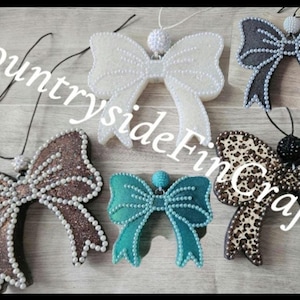 May include: Five bow-shaped ornaments with hanging strings. One is white, one black, one bronze glitter, one teal, and one leopard print. Each bow is outlined with small white or black beads and features a matching beaded sphere at the top center.