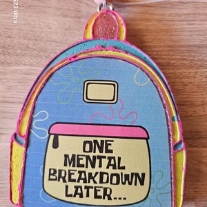May include: A blue, pink, and yellow backpack-shaped charm with the text "ONE MENTAL BREAKDOWN LATER...". The charm has a pink bead and a white string for hanging. The design includes SpongeBob SquarePants-themed elements.