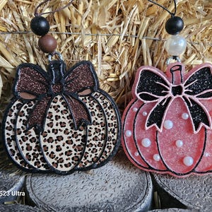 May include: Two pumpkin-shaped ornaments with hanging loops. The left ornament features a brown leopard print pattern with a dark brown glitter bow. The right ornament is pink with white pearl accents and a black glitter bow. Both have bead toppers.