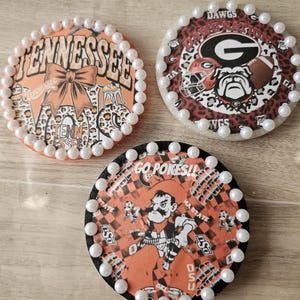 May include: Three round coasters with pearl borders. One features "Tennessee" text and a bow, another has "DAWGS" and a bulldog graphic, and the third says "GO POKES!!" with a cowboy illustration. Each coaster has a unique design.