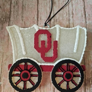 May include: A decorative wagon ornament with a red and black color scheme. The wagon features a red "OU" logo, black wheels with red spokes, and a white top. A black string is attached for hanging.