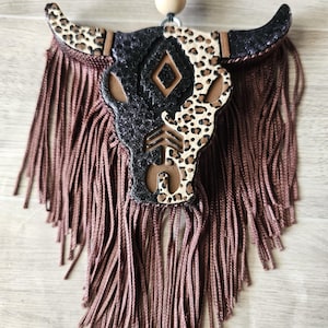 May include: A decorative bull skull ornament featuring a split design with black glitter and a leopard print pattern. The skull includes a central diamond and arrow cutout, accented by long brown fringe hanging from the base and a black and beige bead.