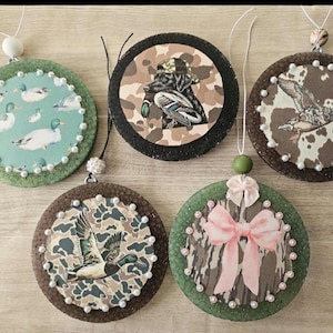 May include: A collection of five round ornaments with duck and hunting-themed designs. Each ornament features a different image, including ducks, a dog, and camouflage patterns. They have beaded edges and hanging strings.